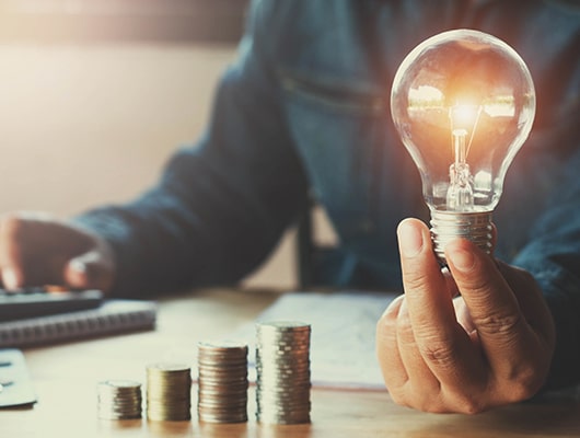 A person holds a lit light bulb next to a staggered stack of coins.