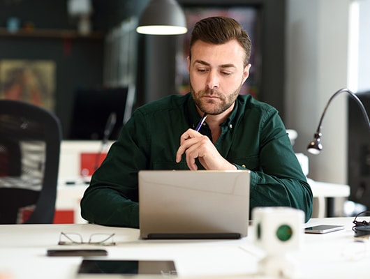 A young, dark haired man, ponders information on a computer screen.